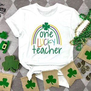Op de afbeelding: Wit T-shirt met een regenboog en klaverontwerp, de tekst luidt "one LUCKY teacher". De afbeelding bevat groene klaverdecoraties, een groene kralenketting en jute banners met klaverontwerpen, perfect voor St. Patrick's Day.