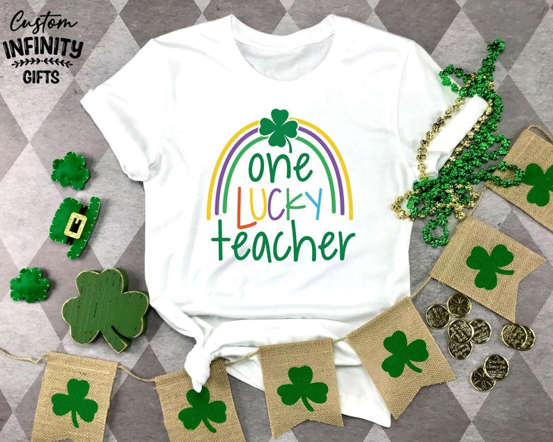 Op de afbeelding: Wit T-shirt met een regenboog en klaverontwerp, de tekst luidt "one LUCKY teacher". De afbeelding bevat groene klaverdecoraties, een groene kralenketting en jute banners met klaverontwerpen, perfect voor St. Patrick's Day.