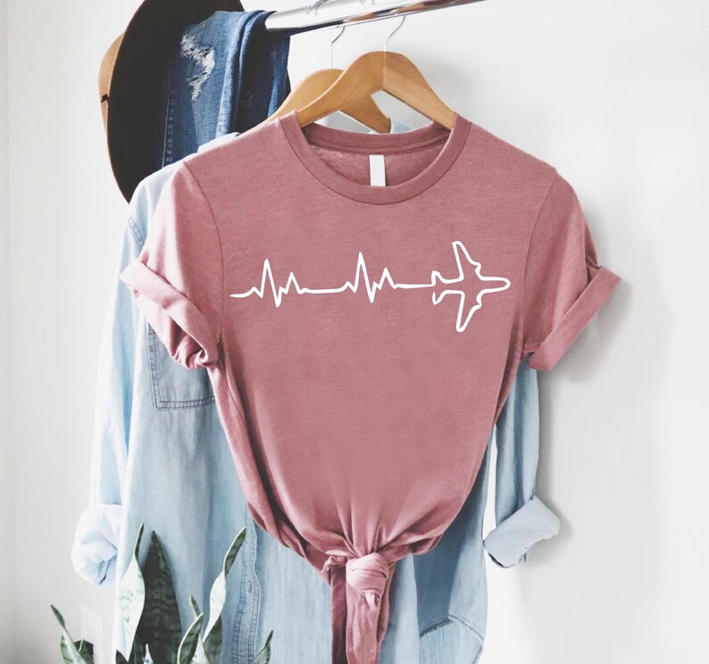 Puede incluir: Camiseta rosa polvoriento con un gr&aacute;fico blanco de una l&iacute;nea de latidos que termina en una silueta de avi&oacute;n. La camiseta de manga corta est&aacute; anudada a la cintura y cuelga de una percha de madera con otras prendas de vestir en el fondo.