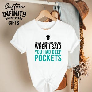 Puede incluir: Camiseta blanca con el texto "I WASN'T COMPLIMENTING YOU WHEN I SAID YOU HAD DEEP POCKETS" en negro y verde azulado. Un gr&aacute;fico de diente negro est&aacute; encima del texto. La camiseta est&aacute; colgada en una percha de madera.