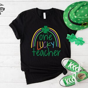 Op de afbeelding: Zwart T-shirt met een regenboogontwerp en de tekst "one lucky teacher". Een groene St. Patrick's Day hoed, klaverbril en groene sneakers staan ook op de foto. Het shirt is een casual stijl.