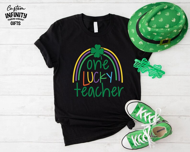 Op de afbeelding: Zwart T-shirt met een regenboogontwerp en de tekst "one lucky teacher". Een groene St. Patrick's Day hoed, klaverbril en groene sneakers staan ook op de foto. Het shirt is een casual stijl.
