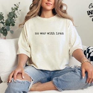 May include: Cream-colored t-shirt with the text "no war with iran" in a simple black font. The shirt is worn with blue jeans. The image also includes the text "Custom Infinity Gifts".