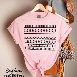 May include: A light pink t-shirt with a black graphic design featuring various line patterns. The shirt is hanging on a wooden hanger. The text "Custom INFINITY GIFTS" is printed on the bottom left of the shirt.