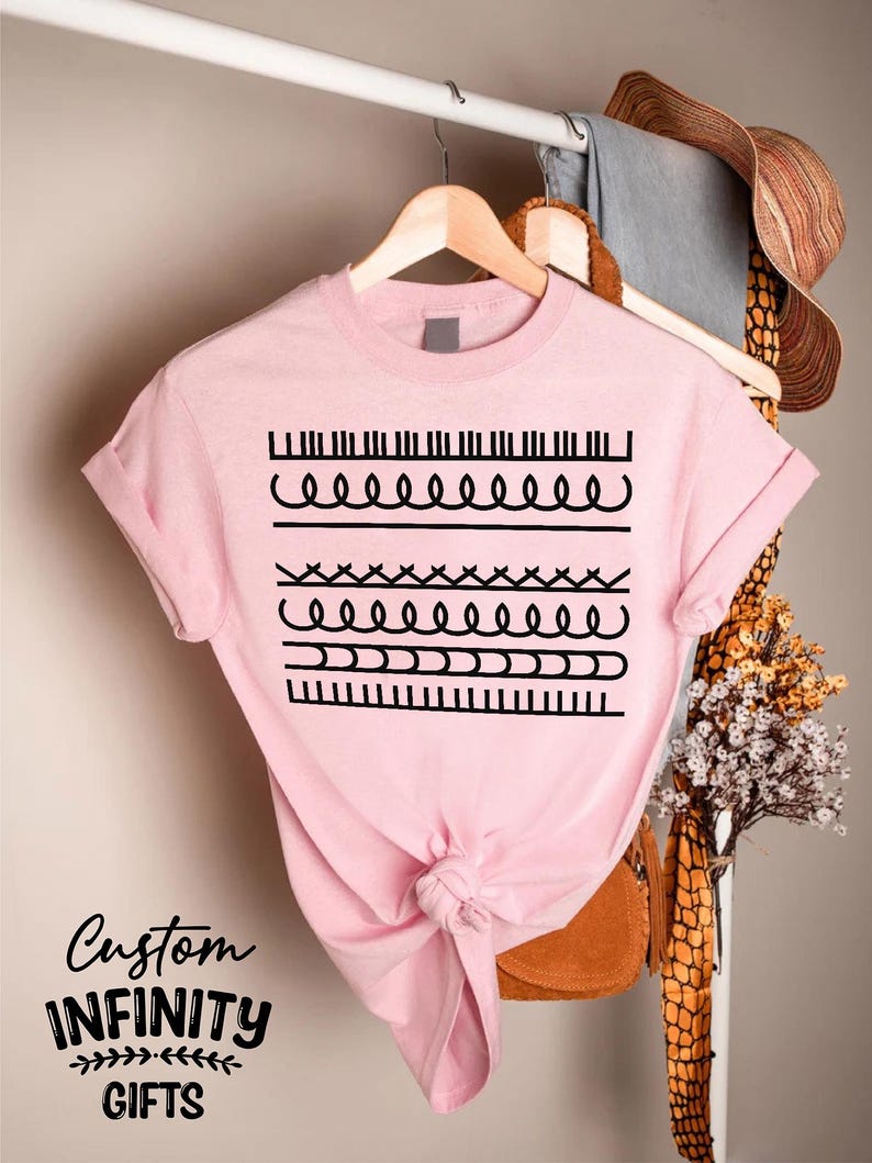 May include: A light pink t-shirt with a black graphic design featuring various line patterns. The shirt is hanging on a wooden hanger. The text "Custom INFINITY GIFTS" is printed on the bottom left of the shirt.