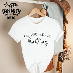 Pu&ograve; includere: T-shirt bianca con la scritta "life is better when I'm knitting" in corsivo nero. La maglietta &egrave; appesa a una gruccia di legno. Le parole "Custom Infinity Gifts" sono in alto a sinistra.