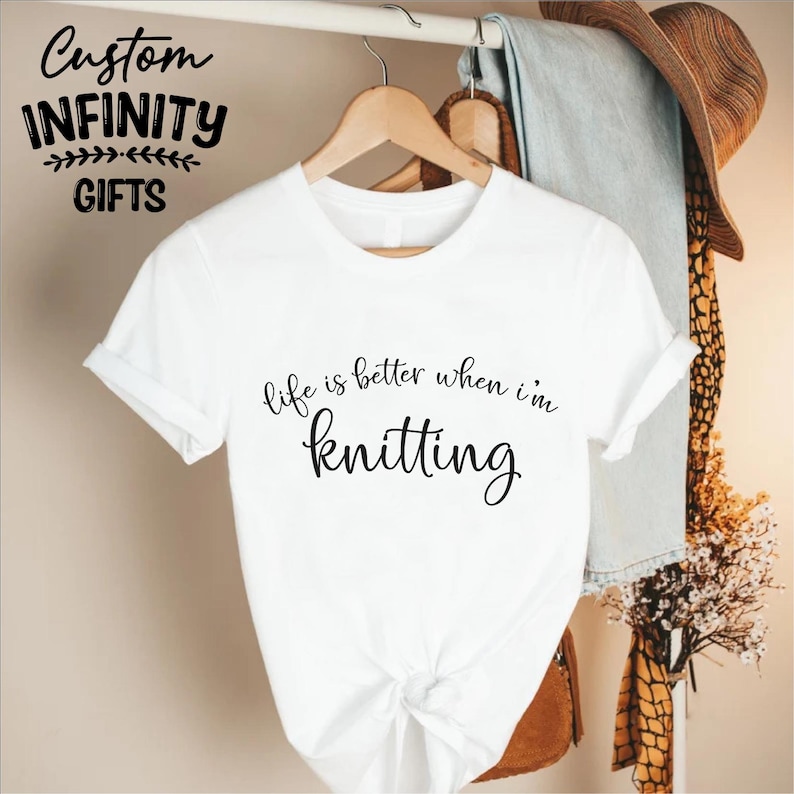 Pu&ograve; includere: T-shirt bianca con la scritta "life is better when I'm knitting" in corsivo nero. La maglietta &egrave; appesa a una gruccia di legno. Le parole "Custom Infinity Gifts" sono in alto a sinistra.