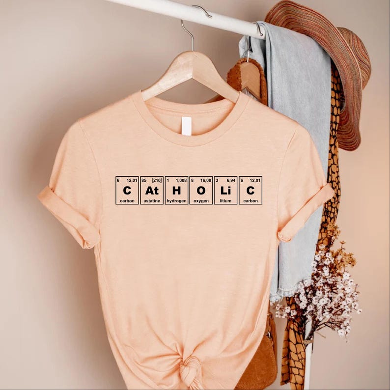 Peut inclure: Un t-shirt p&ecirc;che avec le mot "Catholic" &eacute;pel&eacute; avec les &eacute;l&eacute;ments du tableau p&eacute;riodique. Le t-shirt est suspendu &agrave; un cintre en bois. Le texte comprend carbone, astatine, hydrog&egrave;ne, oxyg&egrave;ne, lithium et carbone.
