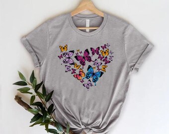 May include: A gray t-shirt with a heart-shaped design made up of colorful butterflies. The butterflies are in shades of pink, blue, orange, and yellow.