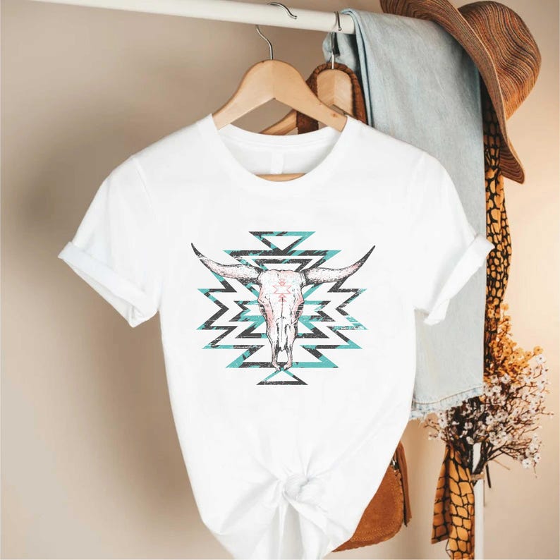 May include: White t-shirt with a turquoise and black geometric design featuring a white bull skull in the center.