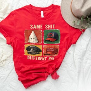May include: Red t-shirt with the text "SAME SHIT DIFFERENT HAT" and images of a KKK hood, a Confederate flag hat, a Nazi hat, and a "Make America Great Again" hat. A beige hat and greenery are in the background.