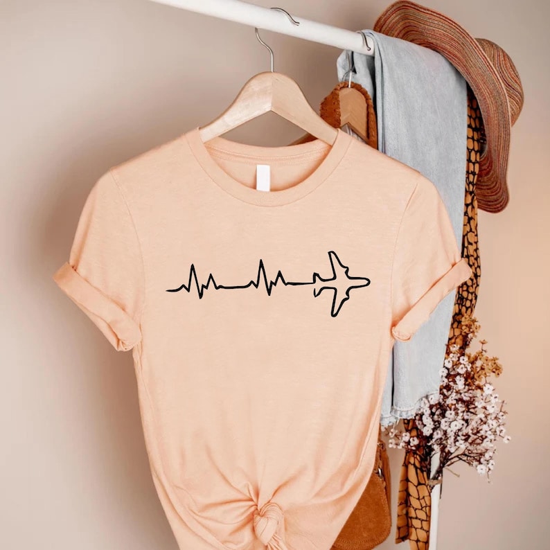 Puede incluir: Camiseta color melocot&oacute;n con un gr&aacute;fico negro de una l&iacute;nea de latidos que se transforma en la silueta de un avi&oacute;n. La camiseta de manga corta est&aacute; colgada en una percha de madera.