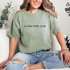 May include: A sage green t-shirt with the text "no war with iran" in black. The shirt is worn with blue jeans. The image also includes the text "Custom Infinity Gifts".