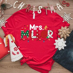 May include: A red t-shirt with the name "Mrs. Miller" in festive, colorful lettering, decorated with Christmas lights and a gingerbread man. The shirt is surrounded by Christmas decorations, including snowflakes and a decorative pillow.