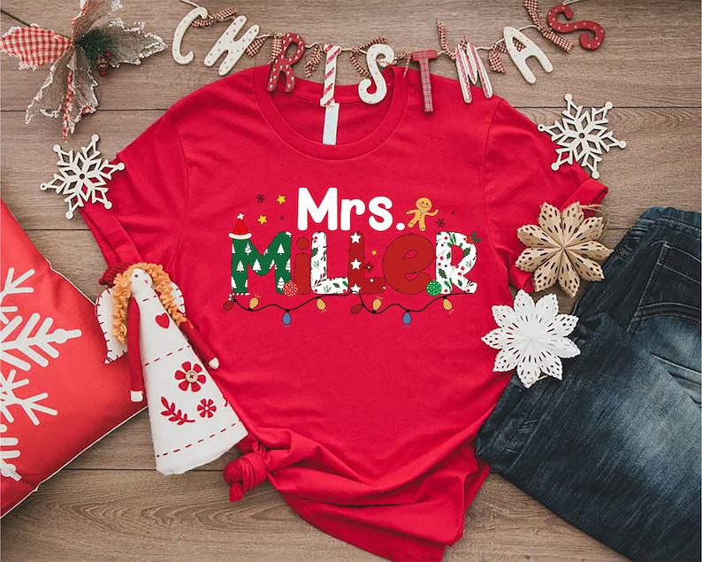 May include: A red t-shirt with the name "Mrs. Miller" in festive, colorful lettering, decorated with Christmas lights and a gingerbread man. The shirt is surrounded by Christmas decorations, including snowflakes and a decorative pillow.