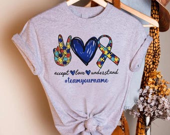 May include: A light gray t-shirt with a peace sign, heart, and ribbon design in blue, yellow, and red puzzle piece patterns. The shirt has the words "accept love understand" and "#teamyourname" in blue.