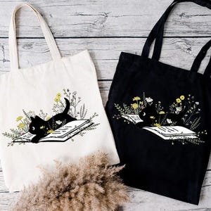 May include: Two tote bags with black cat illustrations. One bag is white with a black cat reading a book surrounded by flowers. The other bag is black with a black cat reading a book surrounded by flowers.