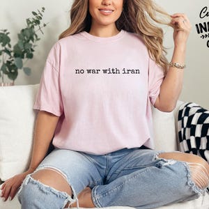 May include: A light pink t-shirt with the text "no war with iran" in black. The shirt is part of a collection of custom infinity gifts. The model is wearing blue jeans.