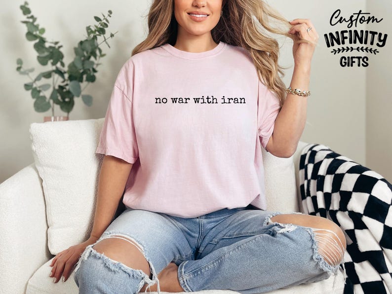 May include: A light pink t-shirt with the text "no war with iran" in black. The shirt is part of a collection of custom infinity gifts. The model is wearing blue jeans.
