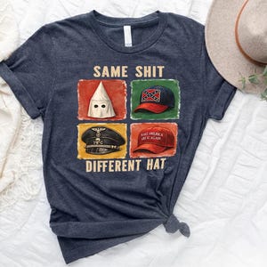 May include: Dark blue t-shirt with the text "SAME SHIT DIFFERENT HAT" and four images of hats. The hats include a KKK hood, a Confederate flag hat, a Nazi hat, and a "Make America Great Again" hat.