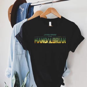 Puede incluir: Camiseta negra con el texto "STAR WARS THE MANDALORIAN" en verde y amarillo. La camiseta est&aacute; colgada en una percha de madera con una camisa vaquera azul claro y un sombrero marr&oacute;n en el fondo.