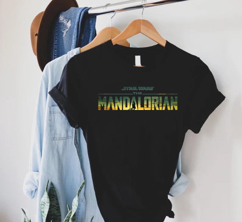 Puede incluir: Camiseta negra con el texto "STAR WARS THE MANDALORIAN" en verde y amarillo. La camiseta est&aacute; colgada en una percha de madera con una camisa vaquera azul claro y un sombrero marr&oacute;n en el fondo.