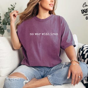 May include: A purple t-shirt with the text "no war with iran" in white. The shirt is worn with blue jeans. The image also shows a black and white checkered blanket and a plant.