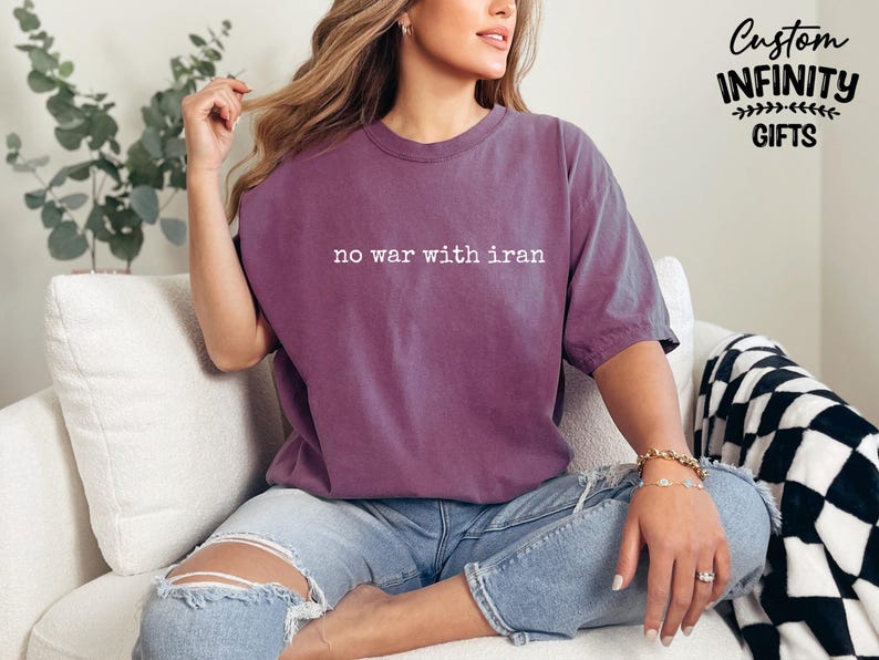 May include: A purple t-shirt with the text "no war with iran" in white. The shirt is worn with blue jeans. The image also shows a black and white checkered blanket and a plant.