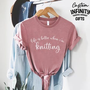 Pu&ograve; includere: Una maglietta rosa polveroso con la scritta bianca "life is better when I'm knitting". La maglietta &egrave; annodata in basso e appesa a una gruccia di legno con altri vestiti. Le parole "Custom Infinity Gifts" sono nell'angolo in alto a destra.