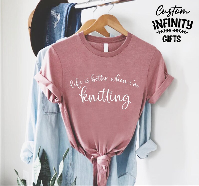 Pu&ograve; includere: Una maglietta rosa polveroso con la scritta bianca "life is better when I'm knitting". La maglietta &egrave; annodata in basso e appesa a una gruccia di legno con altri vestiti. Le parole "Custom Infinity Gifts" sono nell'angolo in alto a destra.
