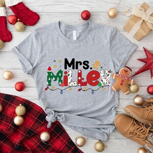 May include: A heather gray t-shirt with the name "Mrs. Miller" in a festive Christmas design. The design includes a Santa hat, gingerbread man, and string lights. Christmas-themed decorations surround the shirt.