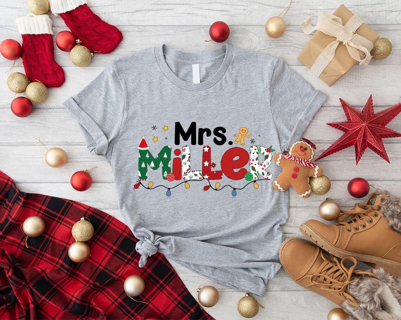 May include: A heather gray t-shirt with the name "Mrs. Miller" in a festive Christmas design. The design includes a Santa hat, gingerbread man, and string lights. Christmas-themed decorations surround the shirt.