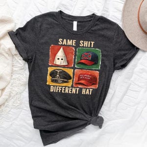May include: Dark gray t-shirt with the text "SAME SHIT DIFFERENT HAT" and images of a KKK hood, a Confederate flag hat, a Nazi hat, and a "Make America Great Again" hat. A tan hat is in the background.