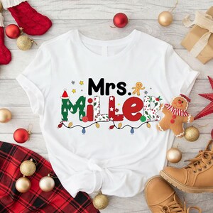 May include: White t-shirt with the text "Mrs. Miller" in a festive Christmas design. The design includes a Santa hat, gingerbread man, and string lights. Christmas ornaments and a pair of boots are in the background.