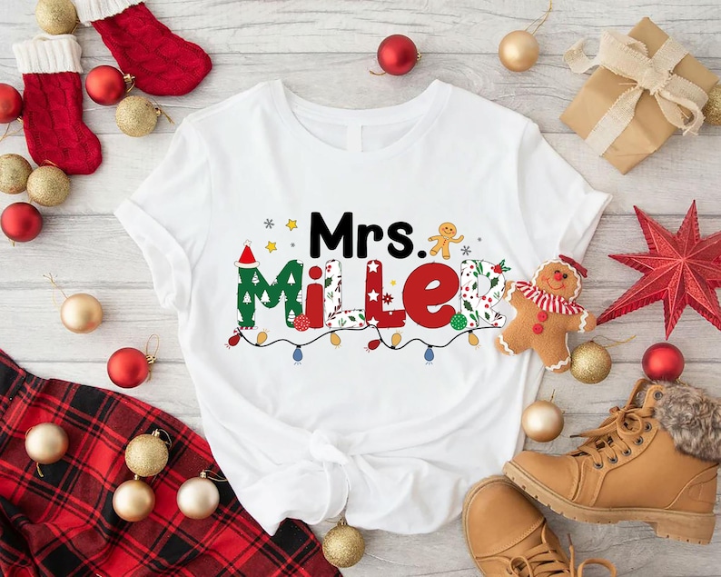 May include: White t-shirt with the text "Mrs. Miller" in a festive Christmas design. The design includes a Santa hat, gingerbread man, and string lights. Christmas ornaments and a pair of boots are in the background.
