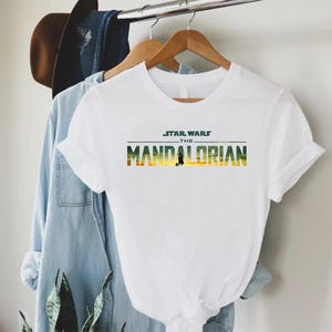 Puede incluir: Camiseta blanca con el texto "STAR WARS THE MANDALORIAN" en verde, amarillo y naranja. La camiseta tiene mangas cortas y cuello redondo. Una camisa vaquera y un sombrero marr&oacute;n cuelgan en el fondo.