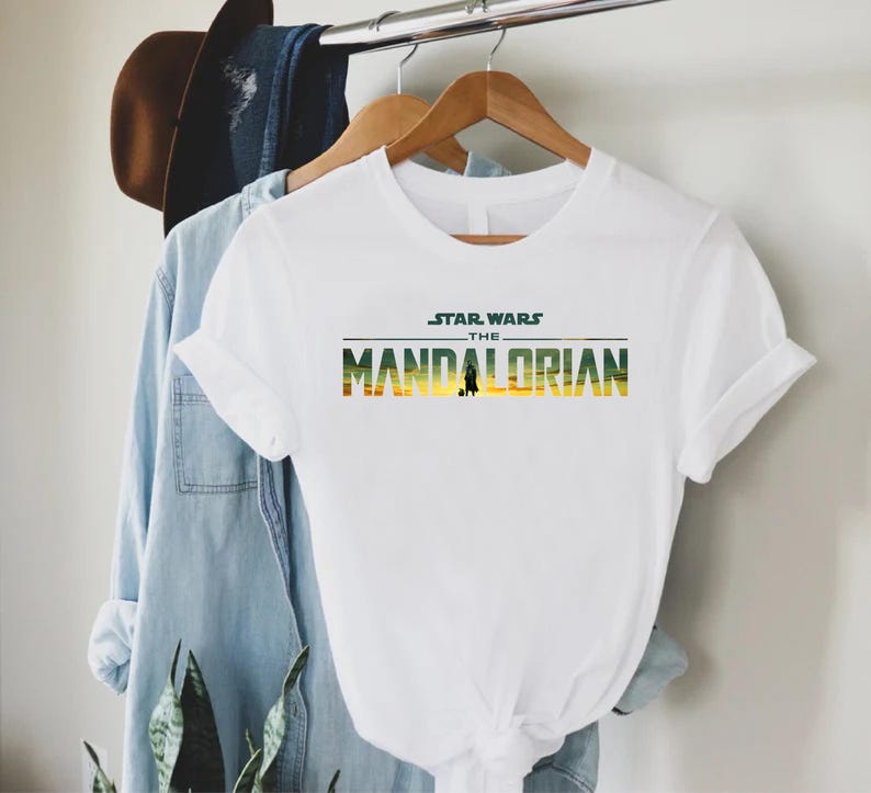 Puede incluir: Camiseta blanca con el texto "STAR WARS THE MANDALORIAN" en verde, amarillo y naranja. La camiseta tiene mangas cortas y cuello redondo. Una camisa vaquera y un sombrero marr&oacute;n cuelgan en el fondo.