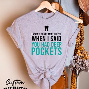 Puede incluir: Una camiseta gris jaspeado con el texto "I WASN'T COMPLIMENTING YOU WHEN I SAID YOU HAD DEEP POCKETS" y un gr&aacute;fico de diente. La camiseta est&aacute; anudada a la cintura y colgada de una percha de madera. Las palabras "Custom Infinity Gifts" tambi&eacute;n est&aacute;n impresas en la camiseta.