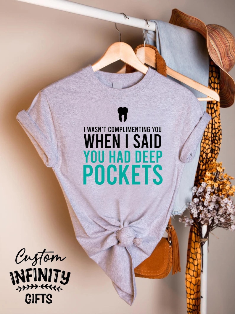 Puede incluir: Una camiseta gris jaspeado con el texto "I WASN'T COMPLIMENTING YOU WHEN I SAID YOU HAD DEEP POCKETS" y un gr&aacute;fico de diente. La camiseta est&aacute; anudada a la cintura y colgada de una percha de madera. Las palabras "Custom Infinity Gifts" tambi&eacute;n est&aacute;n impresas en la camiseta.