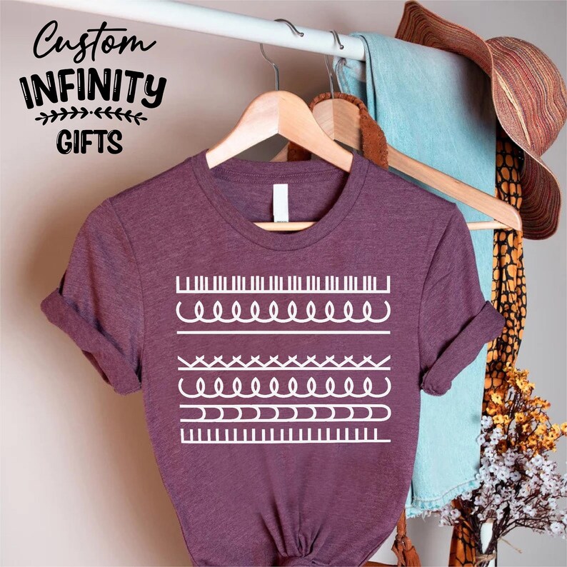 May include: A maroon t-shirt with white graphic designs of various stitch patterns. The shirt is hanging on a wooden hanger. The text "Custom INFINITY GIFTS" is in the upper left corner.
