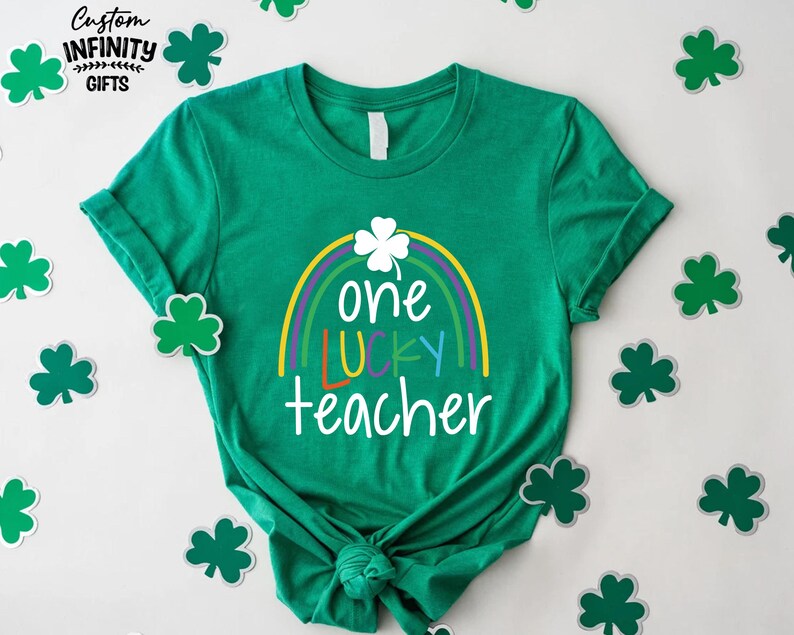 Op de afbeelding: Een groen t-shirt met een regenboog- en klaverontwerp. Het shirt heeft de tekst "one LUCKY teacher" in wit, geel, oranje en blauw. Het shirt is omgeven door klaveruitsparingen.