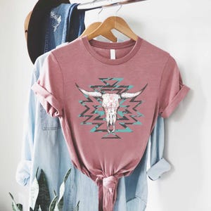 May include: Dusty rose t-shirt featuring a white bull skull graphic with long horns, set against a turquoise and black geometric pattern. The shirt has rolled-up sleeves and is knotted at the waist, suggesting a casual style.