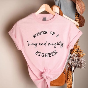 May include: Pink t-shirt with the text "Mother of a Tiny and Mighty Fighter" in black lettering.