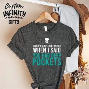 Puede incluir: Camiseta gris oscuro con el texto "I WASN'T COMPLIMENTING YOU WHEN I SAID YOU HAD DEEP POCKETS" en blanco y turquesa. Un gr&aacute;fico de diente blanco est&aacute; encima del texto. La camiseta est&aacute; colgada en una percha de madera.