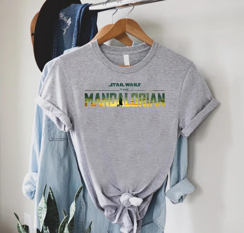 Puede incluir: Una camiseta gris con el texto "STAR WARS THE MANDALORIAN" en verde, amarillo y naranja. La camiseta est&aacute; colgada en una percha de madera, con otras prendas en el fondo.