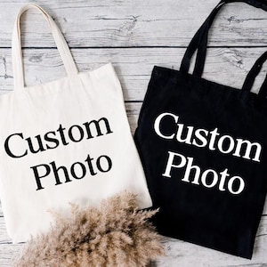 May include: Two tote bags, one cream-colored and one black, with long handles. The words "Custom Photo" are printed in large, bold, white letters on the cream bag and black letters on the black bag. The bags are on a light wood-grain surface.