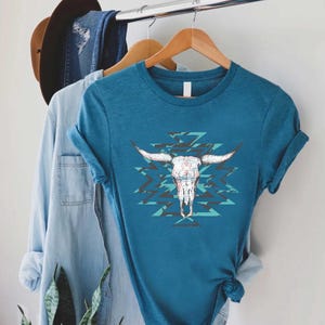May include: Teal-colored t-shirt featuring a white bull skull graphic with long horns and a turquoise and black geometric pattern. The shirt is hanging on a wooden hanger with other clothing items, including a denim shirt and a brown hat.