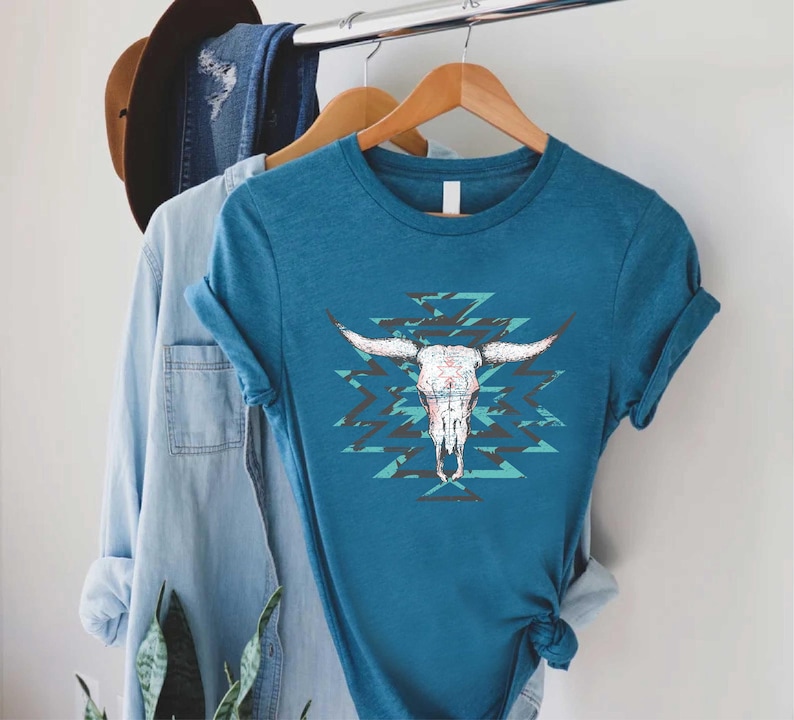 May include: Teal-colored t-shirt featuring a white bull skull graphic with long horns and a turquoise and black geometric pattern. The shirt is hanging on a wooden hanger with other clothing items, including a denim shirt and a brown hat.
