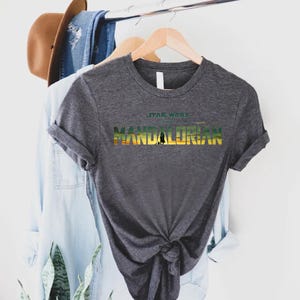 Puede incluir: Camiseta gris oscuro con el texto "STAR WARS THE MANDALORIAN" en un dise&ntilde;o colorido. La camiseta est&aacute; colgada en una percha de madera, con otras prendas y un sombrero en el fondo.