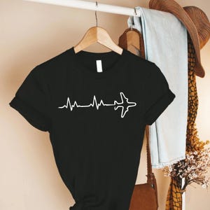 Puede incluir: Camiseta negra con un gr&aacute;fico blanco que combina una l&iacute;nea de latido card&iacute;aco con la silueta de un avi&oacute;n. La camiseta de manga corta est&aacute; colgada en una percha de madera.
